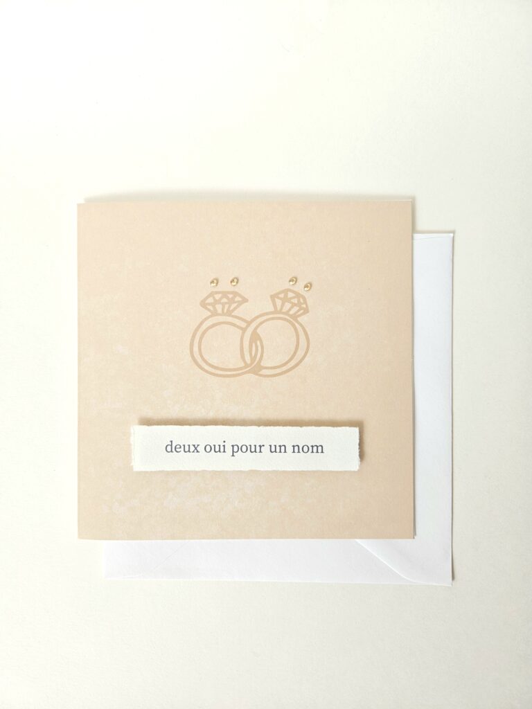 Carte de mariage avec deux alliances entrelacées et le message "deux oui pour un nom", décorée de petites perles.